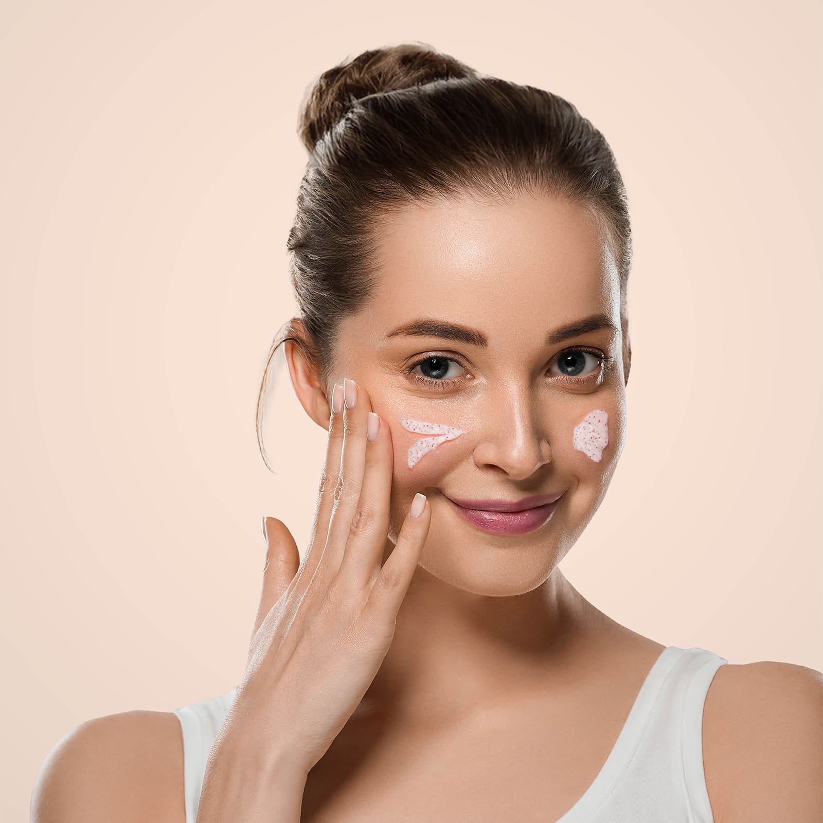 Smiling woman applying pink face cream or scrub to her cheeks, wearing a white tank top against a soft beige background