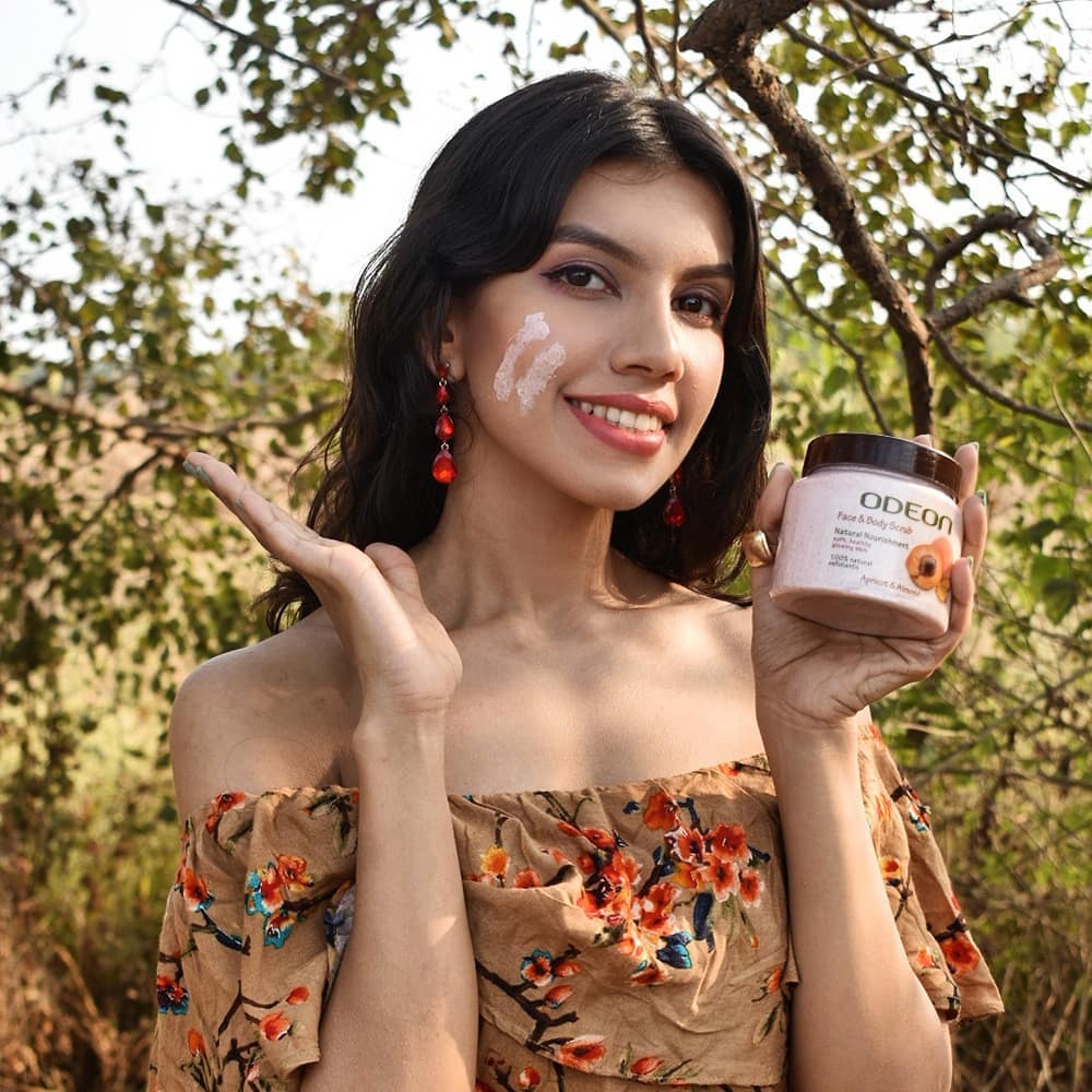 Smiling woman applying Odeon's Apricot & Almond face and body scrub, holding product jar in a natural outdoor setting