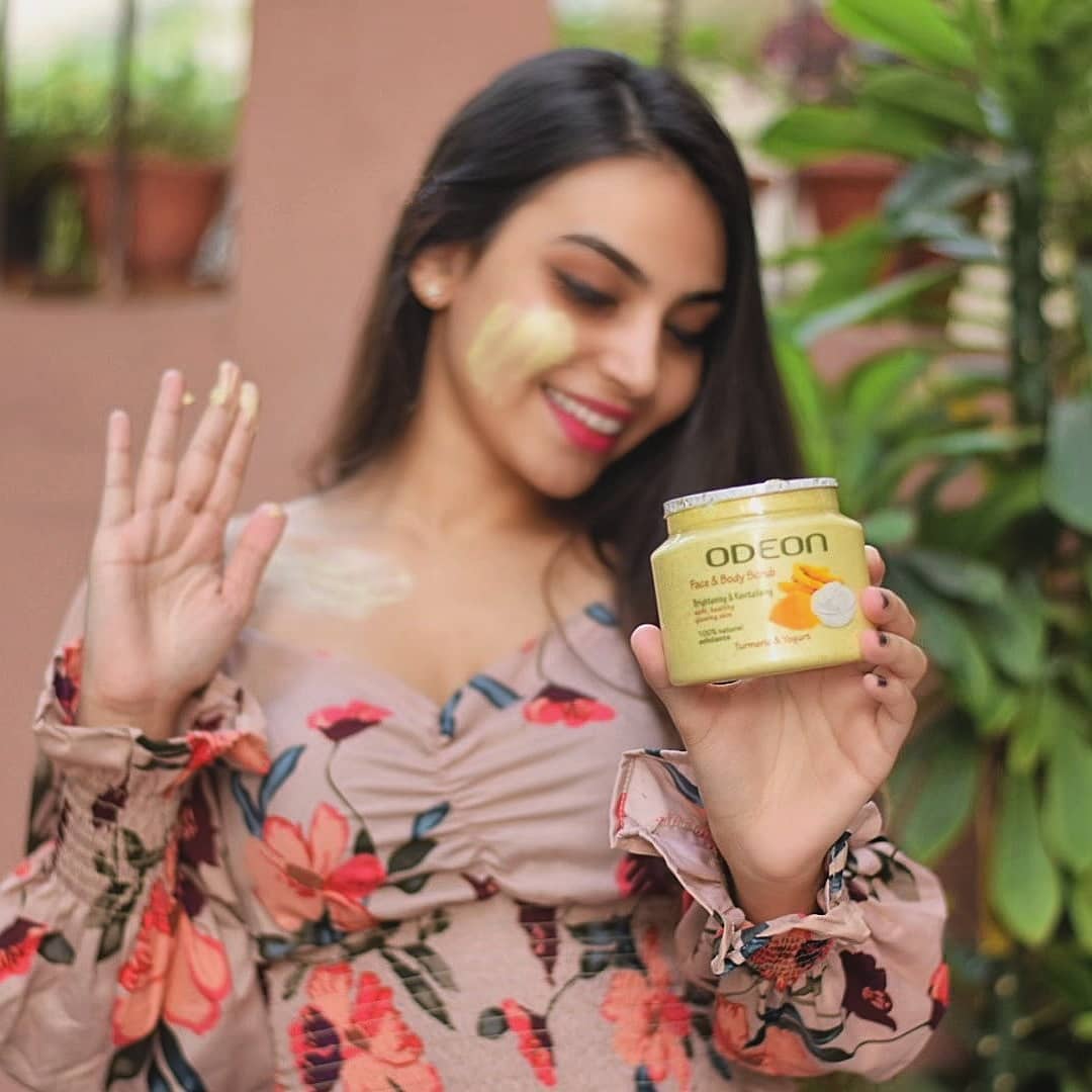 Smiling woman applying skincare product while holding a jar of ODEON Face & Body Scrub with turmeric and yogurt, wearing a floral dress outdoors.