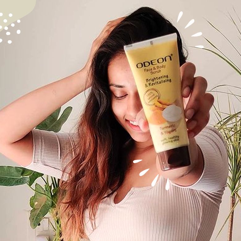 Smiling woman holding up a tube of ODEON Face & Body Scrub with turmeric and yogurt for brightening and revitalizing, standing indoors with plants in the background.