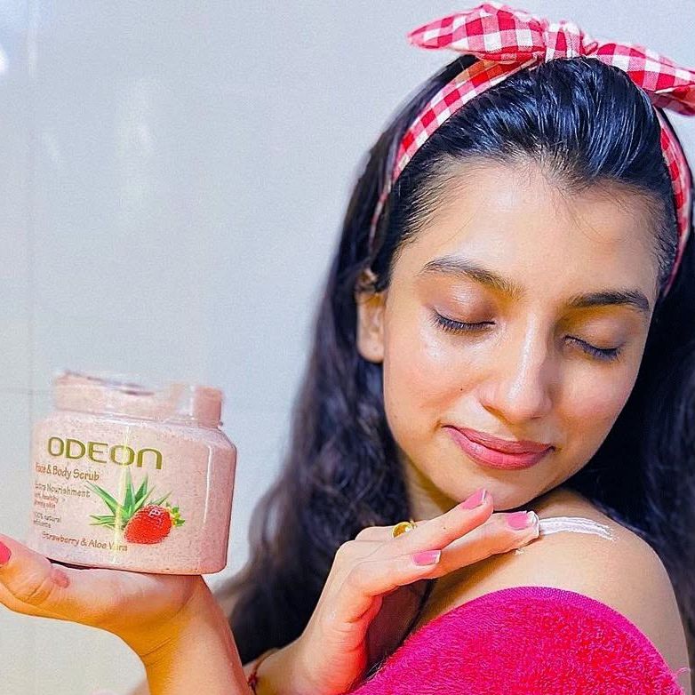 Young woman with a pink headband applying ODEON Face & Body Scrub (Strawberry & Aloe Vera) on her shoulder, smiling softly with closed eyes, showcasing smooth and radiant skin